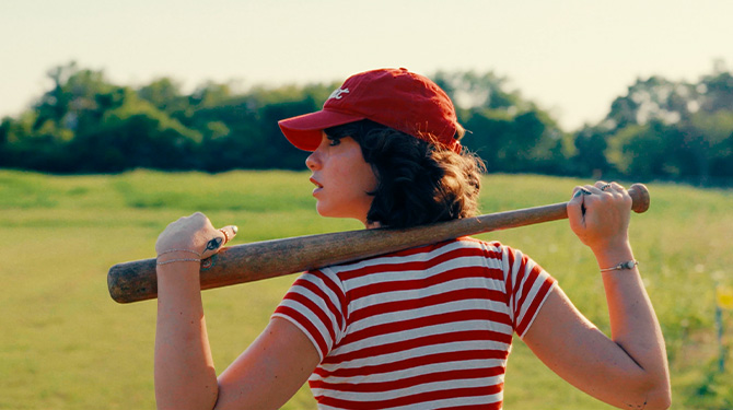 Still from Slugger: a girl in a red cap rests a baseball bat across her shoulders in a sunlit field