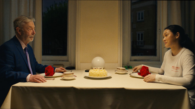 Still from The Unwinding: a widower shares tea and cake with a young woman